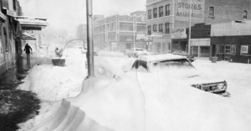 A snowy street scene with a buried car and a person walking, showcasing heavy snowfall and winter conditions.
