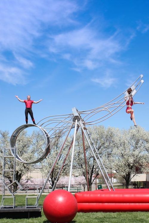 Two performers balance on a large, artistic playground structure against a blue sky and blooming trees.