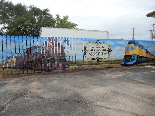 Mural of a train on a fence, featuring the text "Wichita Toy Train Museum" and a colorful train design.