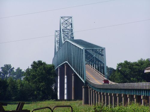 A blue steel bridge spans a river, with a roadway leading up to its elevated section and greenery in the background.