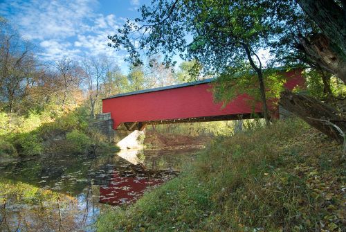A red covered bridge spans a calm river, surrounded by trees and autumn foliage under a blue sky.