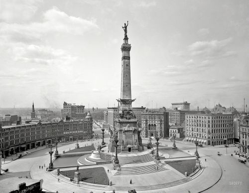 Historic black-and-white photo of a large monument in a city square, surrounded by buildings and people.