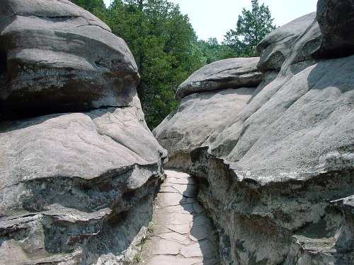 Narrow pathway between large, weathered rock formations surrounded by greenery on a sunny day.