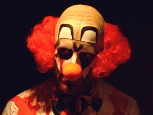 A sinister clown with bright orange hair and a red nose, wearing a painted face and a blood-stained costume.