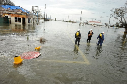 Rescue workers in floodwaters near submerged buildings and a stop sign, with overcast skies in the background.