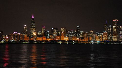 Chicago skyline at night, illuminated buildings reflecting on the water, with colorful lights on skyscrapers.