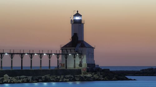 A lighthouse stands on a pier at dusk, with calm waters and a gradient sky transitioning from orange to blue.