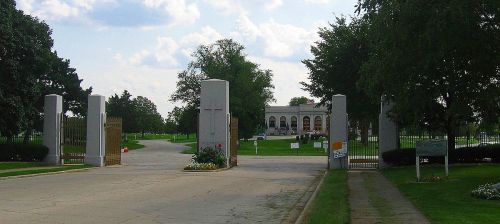 Entrance to a cemetery with large gates, flanked by trees and a building visible in the background under a cloudy sky.