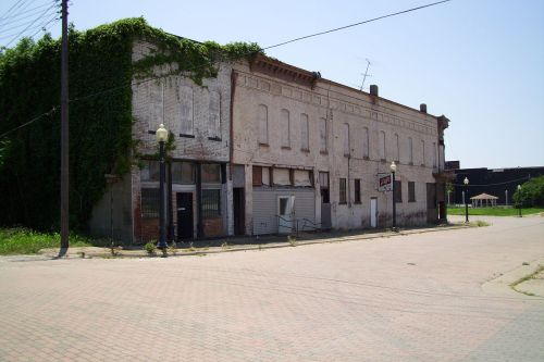 Abandoned brick building with ivy on one side, located at a street corner with a paved road and nearby greenery.