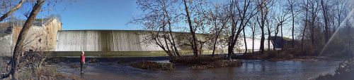 A panoramic view of a dam with a waterfall, surrounded by trees and a flowing stream, under a clear blue sky.