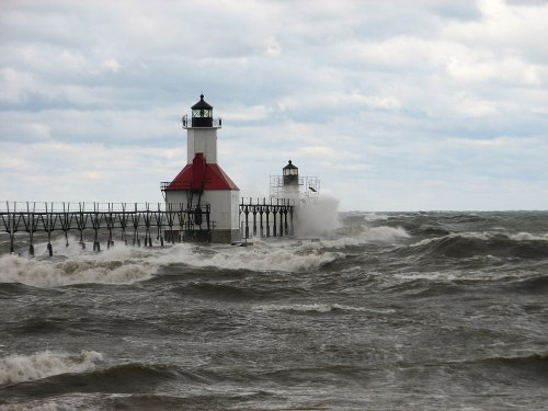 A lighthouse stands on a pier, with waves crashing against it under a cloudy sky.