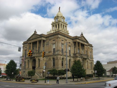 Historic stone courthouse with a clock tower, surrounded by trees and a cloudy sky. Traffic lights visible in front.
