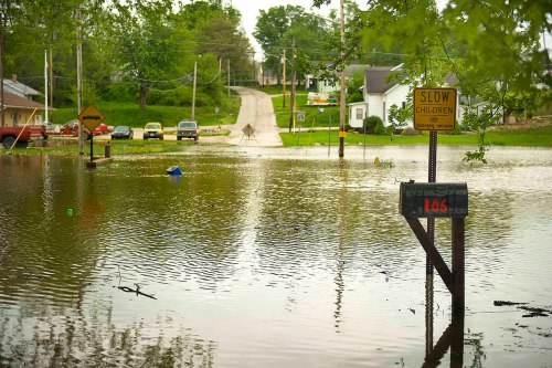 A flooded street with submerged cars and a warning sign, surrounded by trees and a mailbox marked "106."