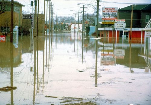 A flooded street with submerged buildings and power lines, reflecting water and debris in a disaster scene.