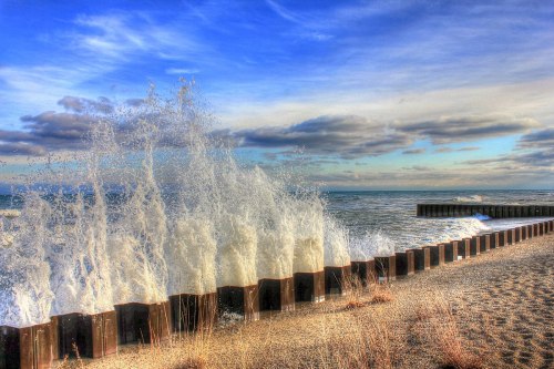 Waves crash against a wooden barrier on a sandy beach under a cloudy blue sky.