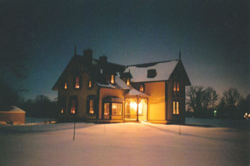 A charming yellow house illuminated at night, surrounded by snow and trees, creating a cozy winter scene.
