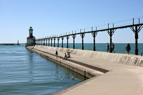 A long pier extends into a calm lake, featuring a lighthouse at the end and people enjoying the sunny day.