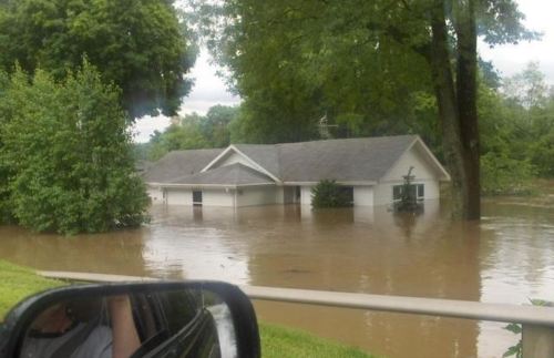 A house partially submerged in floodwaters, surrounded by trees and greenery, with overcast skies above.