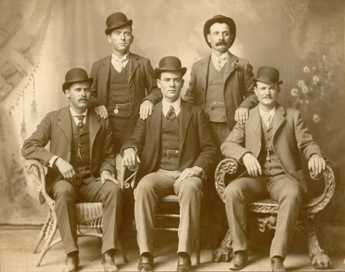 A vintage photo of five men in formal attire and bowler hats, posing together in a studio setting.