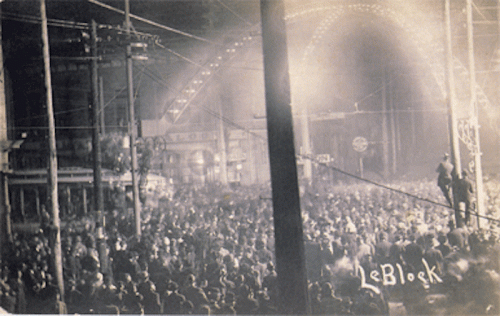 A large crowd gathers at night under illuminated arches, with buildings and power lines in the background.