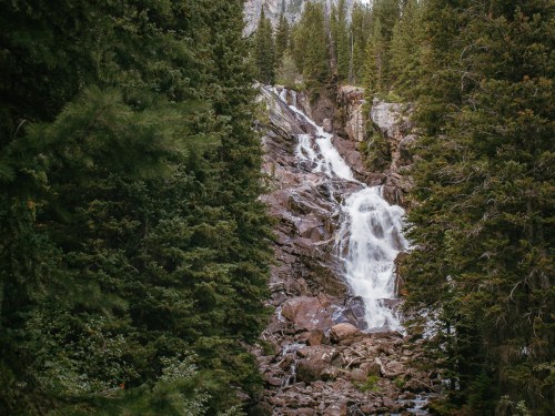 A cascading waterfall flows through rocky terrain, surrounded by lush green trees and mountains in the background.