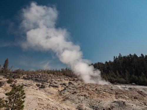 Steam rises from a geothermal area, surrounded by rocky terrain and trees under a clear blue sky.