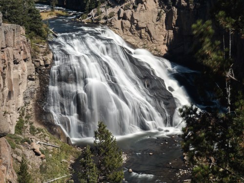 A cascading waterfall flows over rocky cliffs, surrounded by lush greenery and a serene river below.