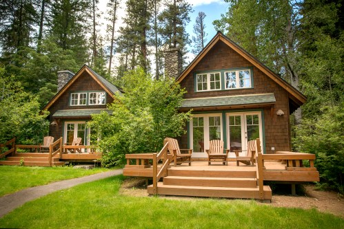 Two cozy cabins with wooden decks surrounded by lush greenery and tall trees under a bright blue sky.