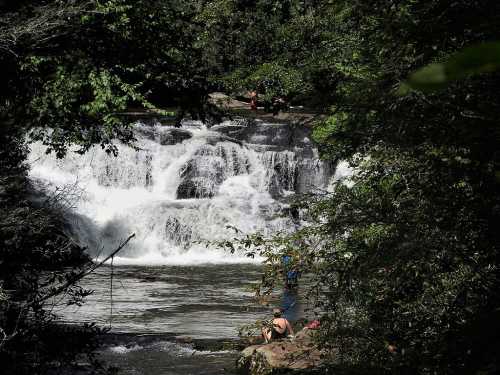 A serene waterfall cascades over rocks, surrounded by lush greenery, with people enjoying the natural setting nearby.