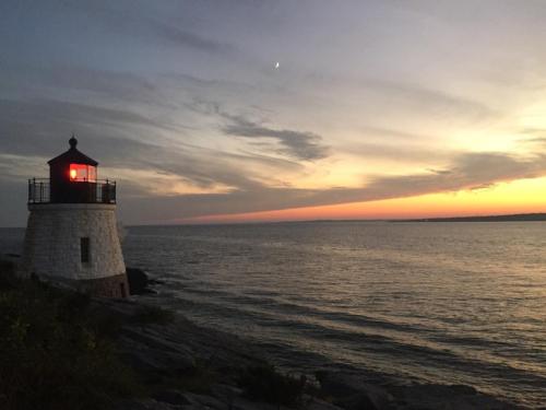 A lighthouse stands on rocky shore at sunset, with a crescent moon visible in the sky above the calm ocean.