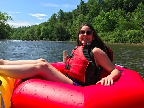 A smiling woman in a red life jacket relaxes on a float in a river, surrounded by lush green trees.
