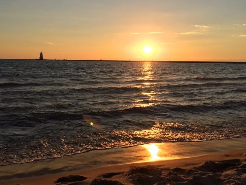 A serene beach scene at sunset, with golden light reflecting on the water and a distant lighthouse silhouette.