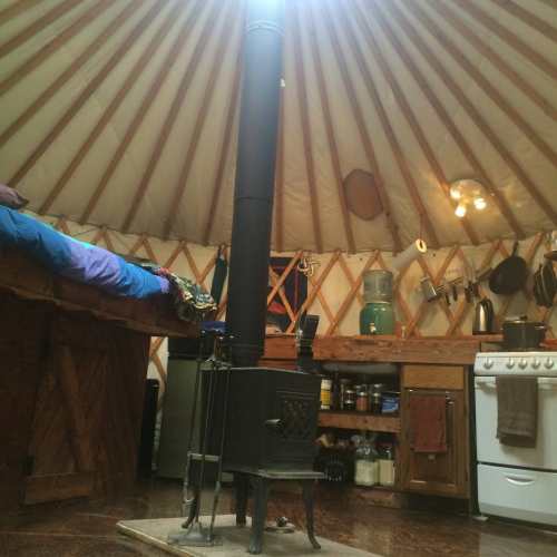 Cozy yurt interior with a wood stove, bed, and kitchen area featuring wooden cabinets and cooking utensils.