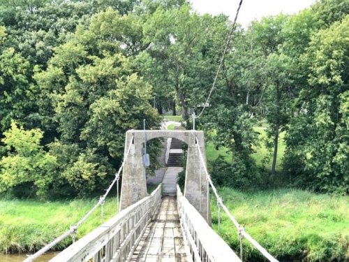 A suspension bridge leads into a lush green park, surrounded by trees and a pathway in the background.