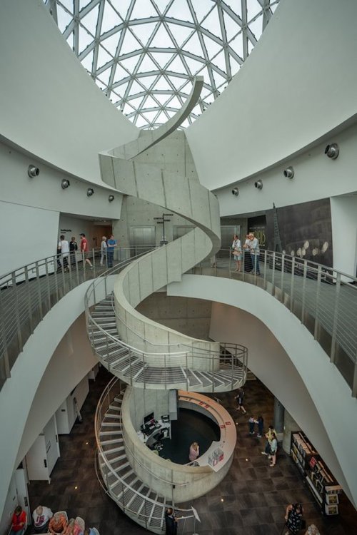 A modern interior featuring a spiral staircase, surrounded by sleek walls and a glass ceiling, with people exploring.