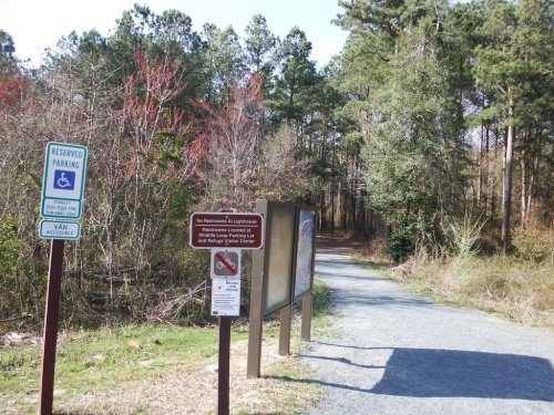 A gravel path leads into a wooded area, with signs for reserved parking and restroom information nearby.