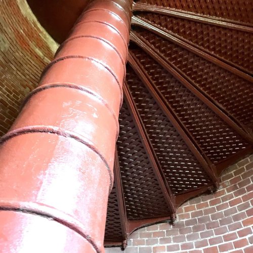 A close-up view of a spiral staircase with a red metal column and textured steps, set against a brick wall.