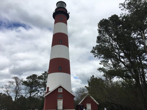 A tall red and white striped lighthouse beside a small building, surrounded by trees under a cloudy sky.
