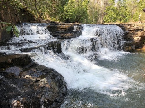 A cascading waterfall flows over rocky steps, surrounded by lush greenery and trees in a serene natural setting.