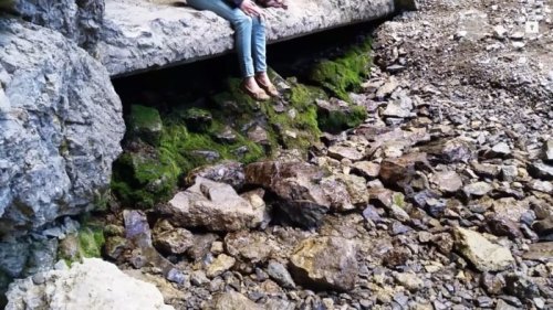 A person sits on a rocky ledge above a stream, surrounded by moss and stones in a natural setting.