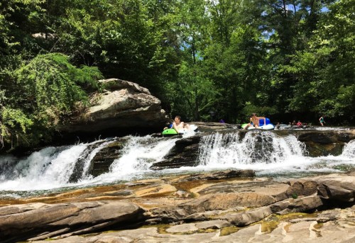 Two people float on inflatable tubes in a river near a small waterfall, surrounded by lush green trees.