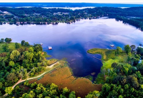 Aerial view of a serene lake surrounded by lush greenery and small boats, with distant hills under a clear sky.