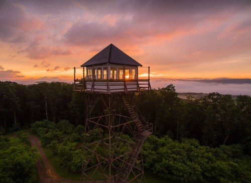 A tall wooden observation tower at sunset, surrounded by lush greenery and misty hills in the background.