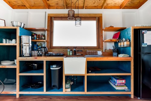 A modern kitchen with wooden shelves, a sink, and various kitchenware, featuring a large window for natural light.