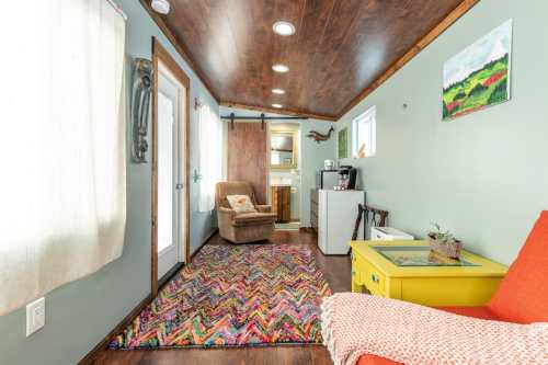 Cozy interior of a tiny home featuring a colorful rug, armchair, and kitchenette with natural light from windows.