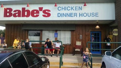 Exterior of Babe's Chicken Dinner House, featuring a wooden facade, sign, and people waiting outside.
