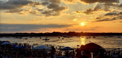 A vibrant sunset over a crowded beach with boats on the water and people enjoying the evening.