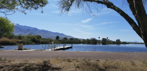 A serene lake scene with a wooden dock, surrounded by trees and mountains under a clear blue sky.