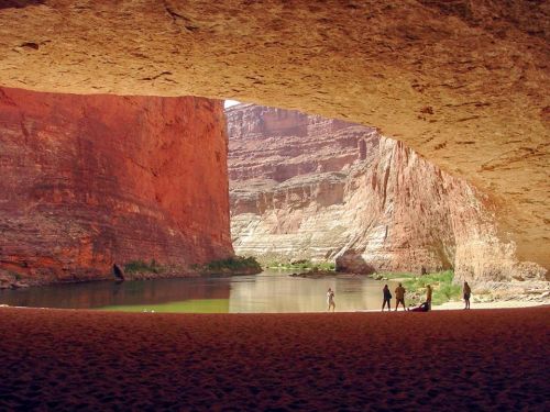 A view from under a rock overhang, showcasing a river and people silhouetted against red canyon walls.