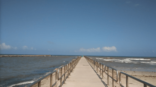 A long pier extends into the ocean under a clear blue sky, with gentle waves lapping at the sides.